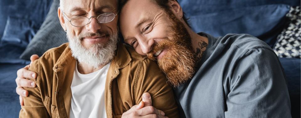 A smiling person with a red beard leans their head on the shoulder of a smiling person with a white beard and glasses.
